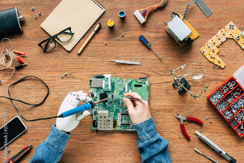 cropped image of electronic engineer with prosthetic arm fixing motherboard by soldering iron and tweezers photo