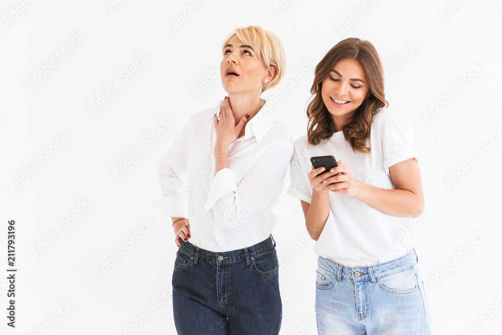 Portrait of caucasian woman 40s looking upward with disinterest while young teen girl holding and using black smartphone, standing isolated over white background Portrait of caucasian woman 40s looking upward with disinterest while young teen girl holding and using black smartphone, standing isolated over white background