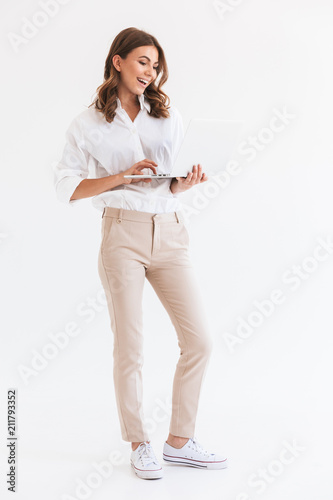 Portrait of adorable smiling woman with long brown hair holding and looking at silver laptop, isolated over white background in studio