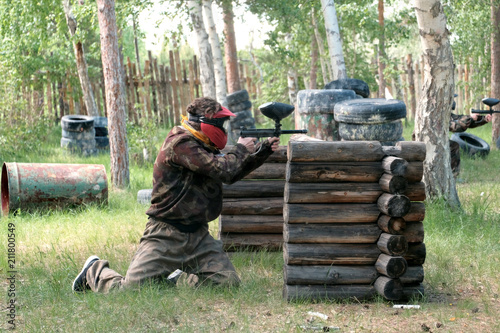 A guy or girl in a protective suit peeks out from behind a wooden shelter during a game of paintball. Landfill in the woods for extreme sports.
