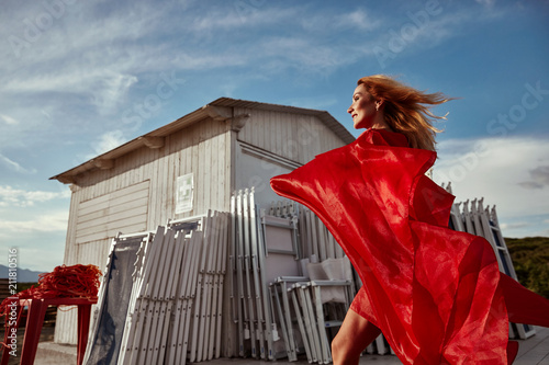 Dynamic outdoor portrait of attractive blonde woman in red flying dress, walking near white wooden building on the shore photo