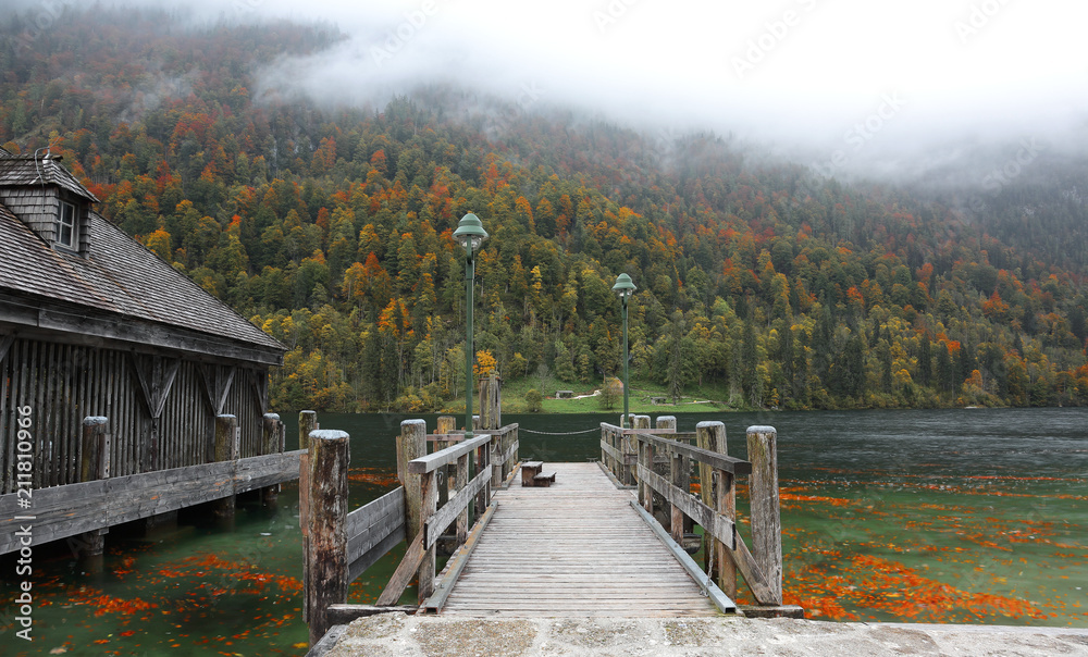 Fototapeta premium An empty wooden pier by beautiful lakeside on a misty foggy morning ~ Gloomy autumn & Beautiful autumn scenery of Koenigssee (King's lake) in Bavaria, Germany