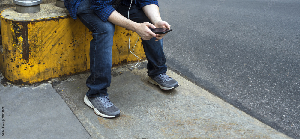 man with jeans and sneakers sitting on a yellow metal and cement blocker playing with his mobile device man with jeans and sneakers sitting on a yellow metal and cement blocker playing with his mobile device