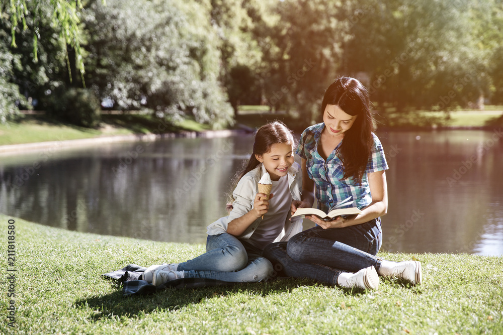 Young woman is sitting together with her daughter and reading a book. They are looking at book together. Girl is leaning to her mom. She is eating ice cream.