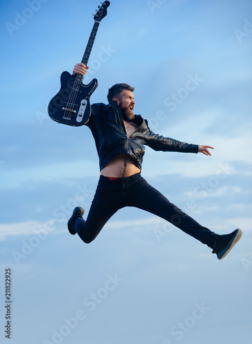 Motion and fun concept. Happy young man jumping in air and playing guitar over blue sky and clouds background