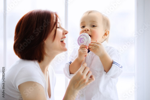 Little boy eating pink and white candy on stick  given by his laughing mother. Happy childhood and family fun concept