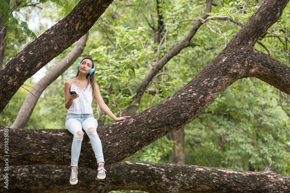 Asian Beautiful happy  woman sit on big tree  and listen to streaming music from smartphone by earbuds. Brunette girl relax in summer park.