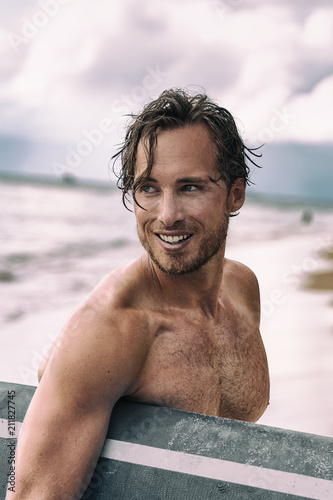 Sexy young man surfer beach lifestyle. Smiling male beauty model tanned relaxing holding surf board on tropical vacation. Healthy skin care portrait concept.