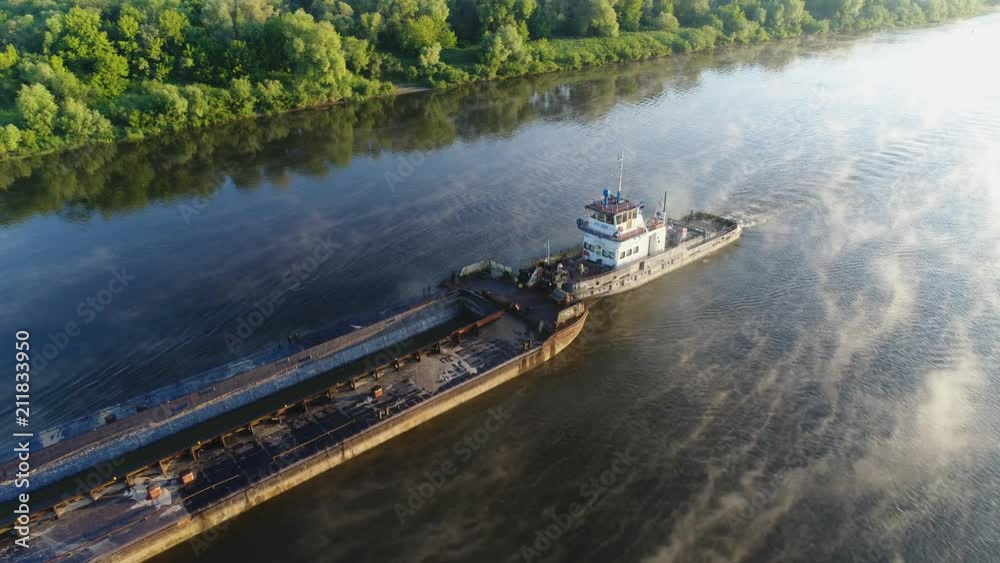 Aerial view. An old tug pushes a barge down a small river at dawn slowly. Thick fog beautifully spreads and reflected from the smooth surface of the water. Transport ship work on the river. Aerial view. An old tug pushes a barge down a small river at dawn slowly. Thick fog beautifully spreads and reflected from the smooth surface of the water. Transport ship work on the river.