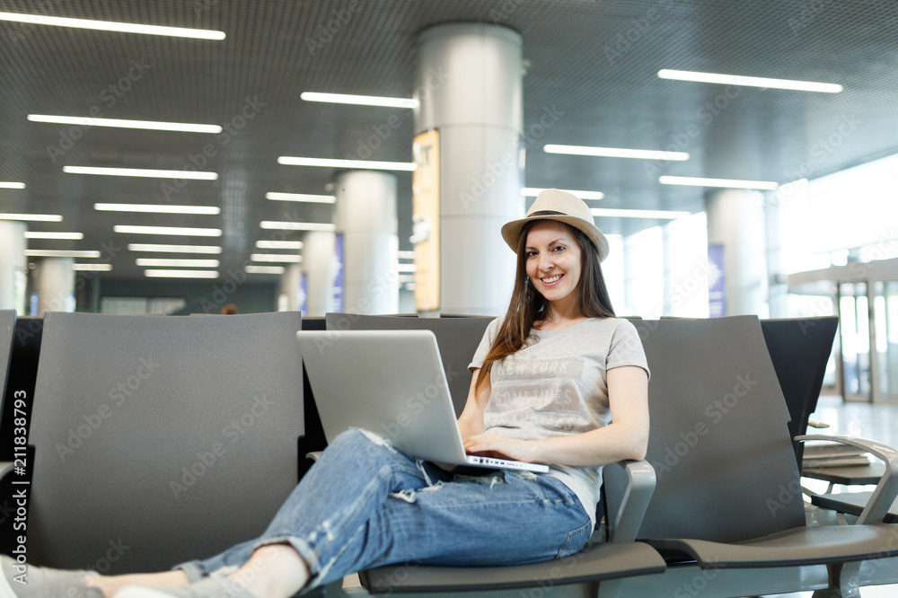 Young smiling traveler tourist woman in hat working on laptop while waiting in lobby hall at international airport. Passenger traveling abroad on weekends getaway. Air travel, flight journey concept. Young smiling traveler tourist woman in hat working on laptop while waiting in lobby hall at international airport. Passenger traveling abroad on weekends getaway. Air travel, flight journey concept.