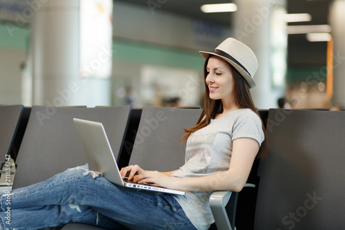 Young relaxed traveler tourist woman sitting working on laptop while waiting in lobby hall at international airport. Passenger traveling abroad on weekends getaway. Air travel, flight journey concept.