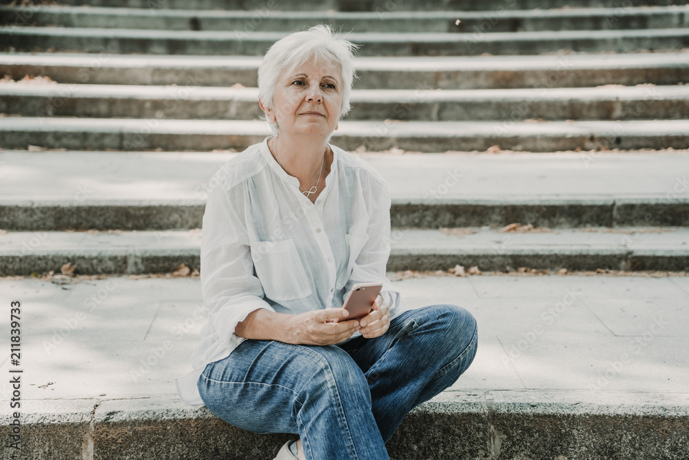 Sweet relaxed middle-aged woman with white hair enjoying a nice spring day in a central park in Madrid. Using her smart phone, talking and sending messages. Lifestyle Sweet relaxed middle-aged woman with white hair enjoying a nice spring day in a central park in Madrid. Using her smart phone, talking and sending messages. Lifestyle