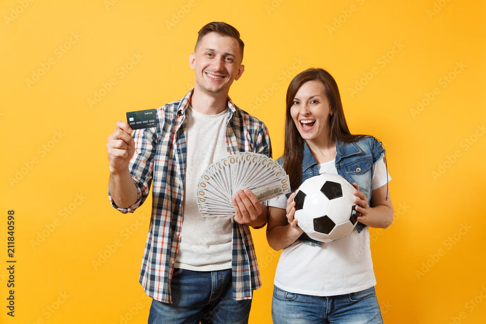 Young win couple, woman man, football fans holding bundle of dollars money, credit card, soccer ball, cheer up support team isolated on yellow background. Sport bet, ardor family lifestyle concept. Young win couple, woman man, football fans holding bundle of dollars money, credit card, soccer ball, cheer up support team isolated on yellow background. Sport bet, ardor family lifestyle concept.