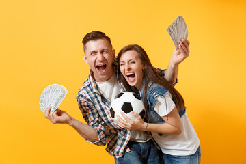 Young win couple, woman man, football fans holding bundle of dollars, cash money, soccer ball, cheer up support team isolated on yellow background. Sport bet, excitement ardor family lifestyle concept