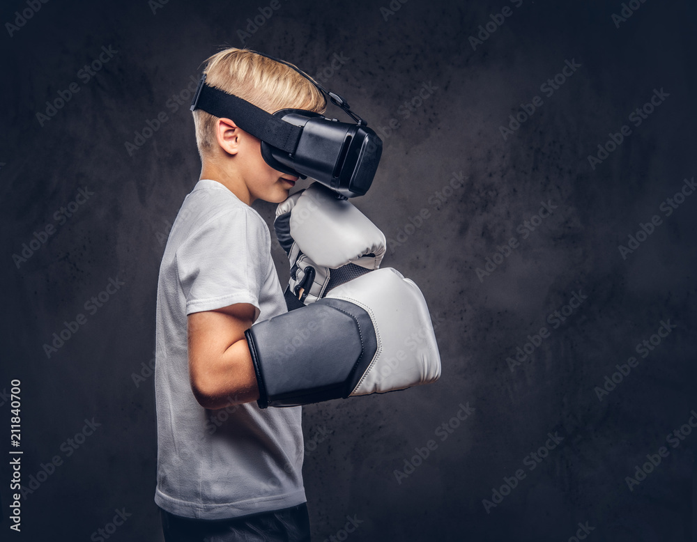Schoolboy boxer with blonde hair dressed in a white t-shirt wearing visual reality glasses and boxing gloves, workout in a studio. Isolated on dark textured background. Schoolboy boxer with blonde hair dressed in a white t-shirt wearing visual reality glasses and boxing gloves, workout in a studio. Isolated on dark textured background.