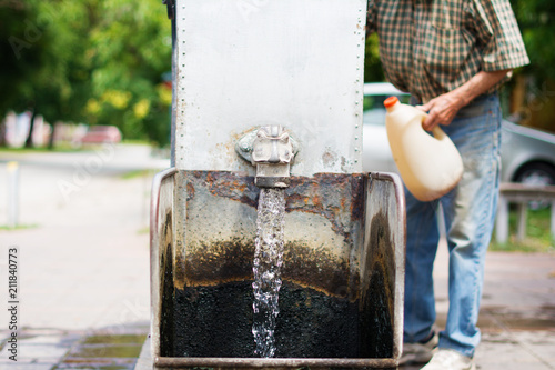 Gothic spigot installed on a flowing artesian wellhead. Selective focus. The blurry contours of a partially visible man holding a plastic demijohn, in the nearest surroundings. photo