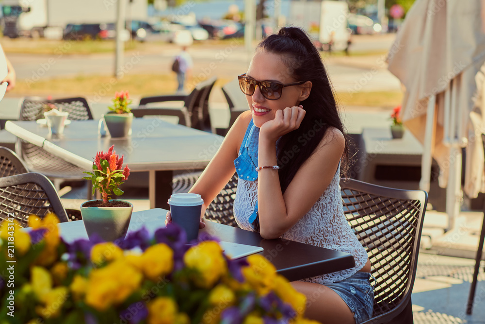 Happy charming brunette girl wearing trendy clothes is enjoying summer day while sitting on a terrace in an outdoor cafe.
