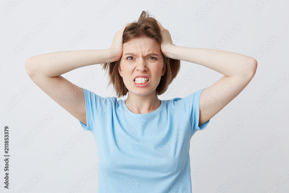 Portrait of angry irritated young woman in blue tshirt standing with hands on head and feels crazy isolated over white background Looks directly in camera Portrait of angry irritated young woman in blue tshirt standing with hands on head and feels crazy isolated over white background Looks directly in camera