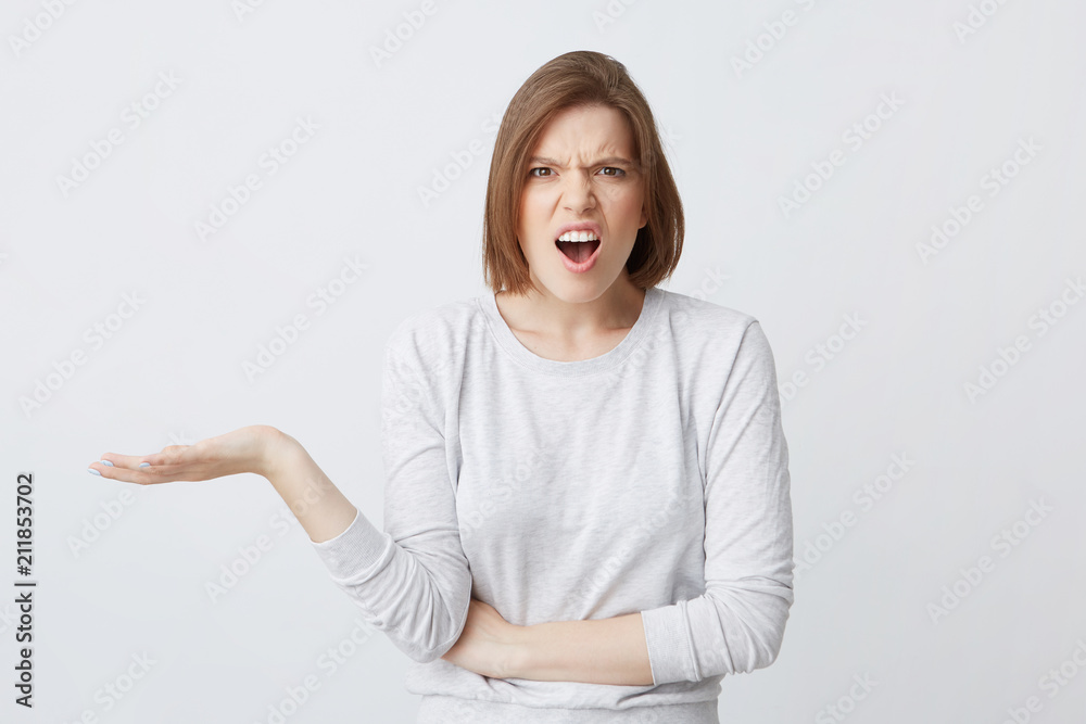 Portrait of angry shocked young woman in longsleeve with opened mouth standing and holding copyspace on palm isolated over white background Portrait of angry shocked young woman in longsleeve with opened mouth standing and holding copyspace on palm isolated over white background