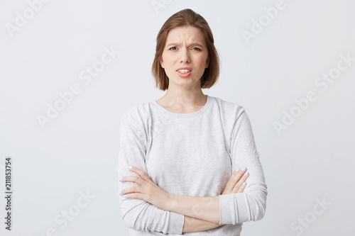 Closeup of angry annoyed young woman in longsleeve standing with arms crossed and feels irritated isolated over white backgound Looks directly in camera photo