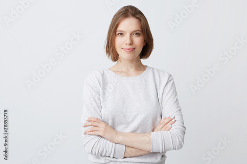 Closeup of happy pretty young woman in longsleeve standing with hands folded and looking directly in camera isolated over white wall Feeling confident photo
