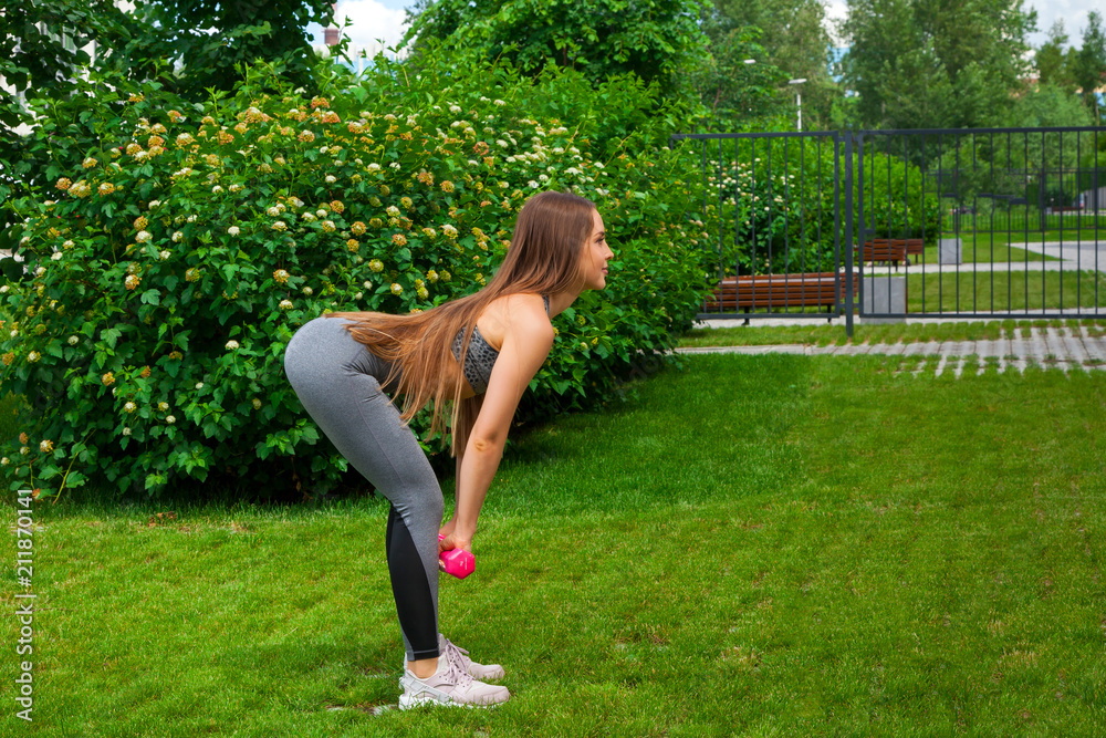 Fototapeta premium A dark-haired woman coach in a sporty short top and gym leggings makes a deadlift with dumbbells on a summer day in a park on a green lawn