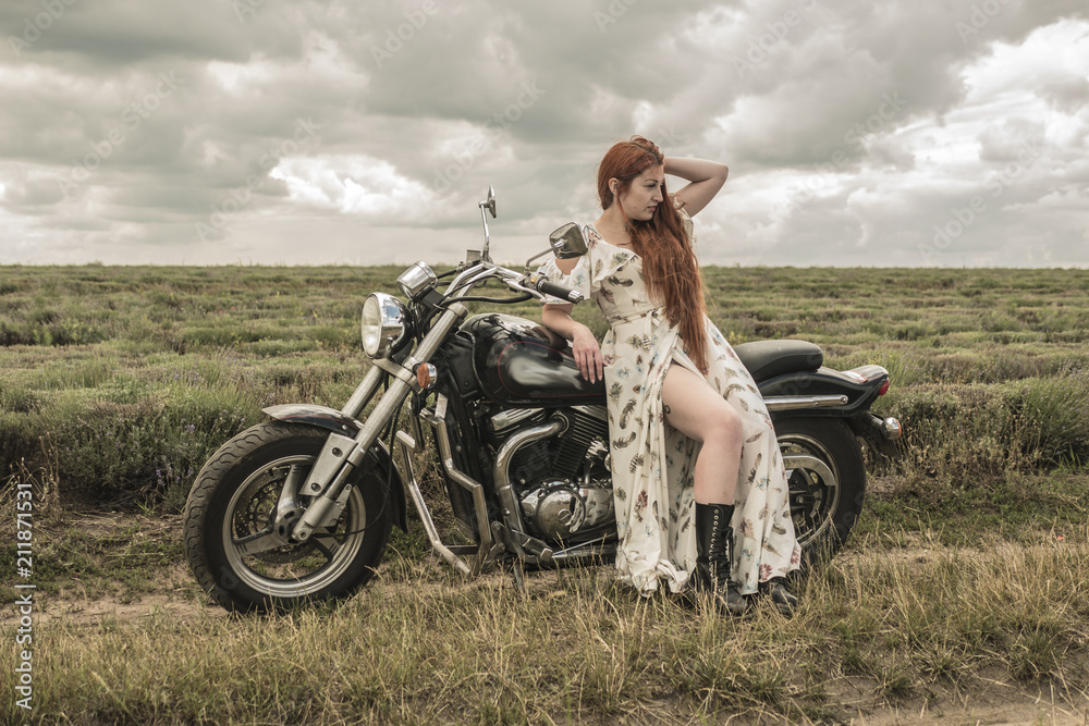 Fototapeta premium red-haired girl in a white dress and boots along with a motorcycle. lavender field