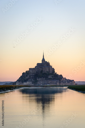 The silhouette of the famous Mont Saint-Michel tidal island in Normandy, France, at sunrise and high tide, reflecting in the still waters of the Couesnon river with the warm colors of the sky. © olrat