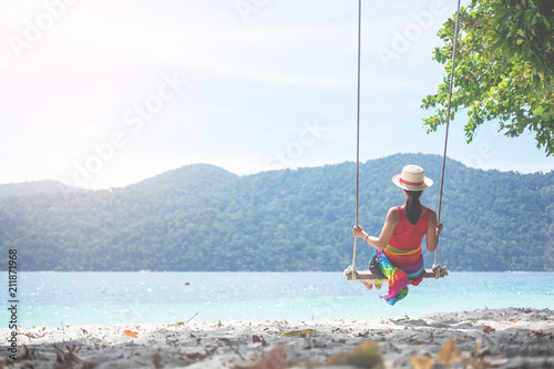 Outdoors lifestyle fashion portrait stunning young girl enjoying on swing on the tropical island. In the background the sea. Wearing stylish dress.