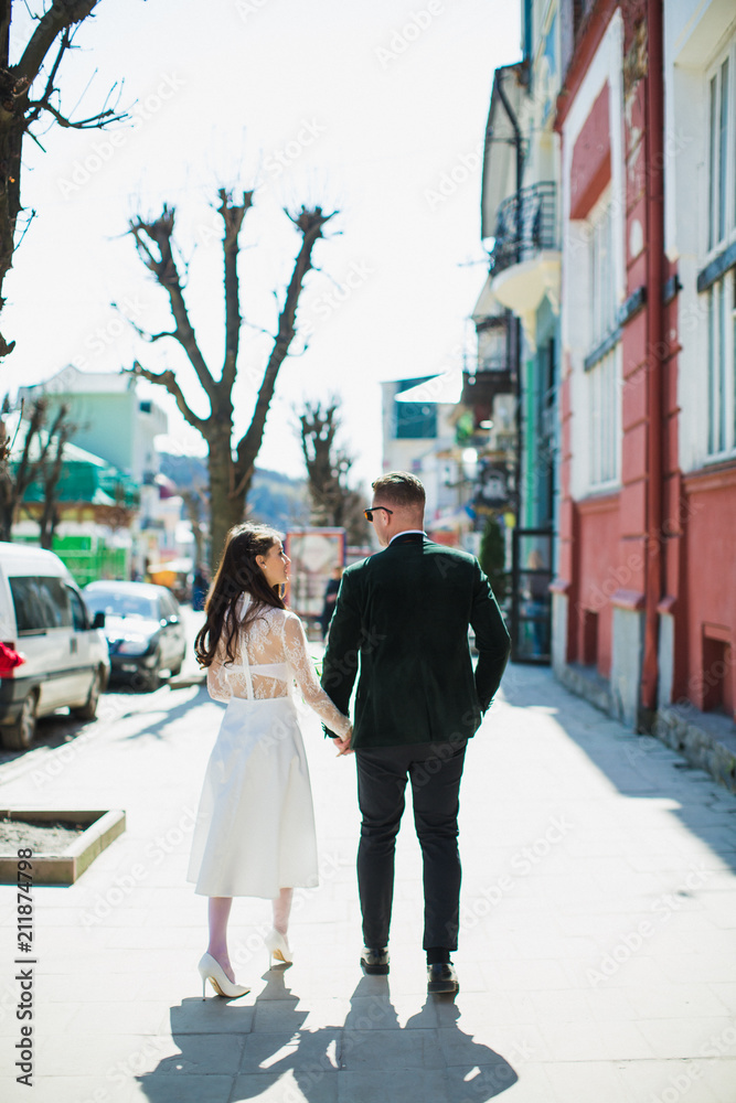 A side view of romantic young stylish newlyweds who are walking and holding their hands outdoors