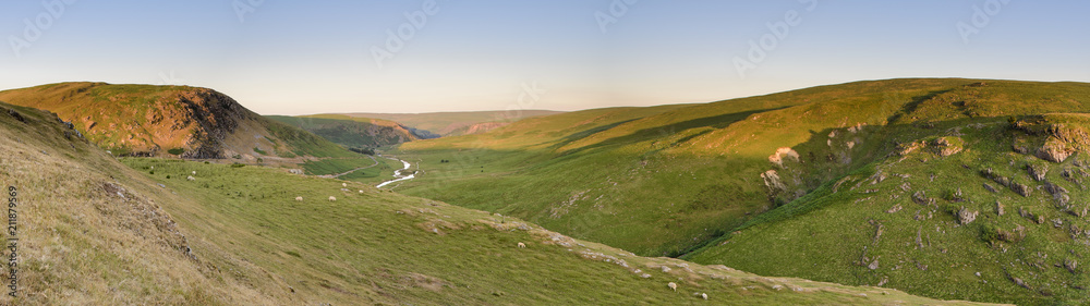 Fototapeta premium Panoramic view of the spectacular Elan Valley, in Powys, mid Wales. Sheep are grazing in the landscape and a small stream runs through the valley. The setting sun is lighting the mountain tops