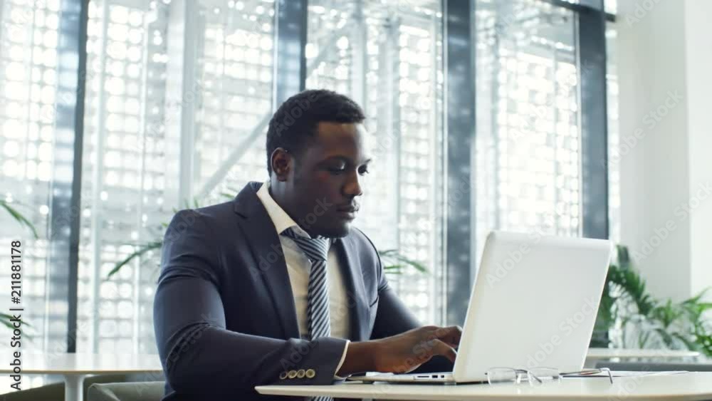 PAN shot of African American businessman sitting at table in modern coworking space and typing on laptop PAN shot of African American businessman sitting at table in modern coworking space and typing on laptop