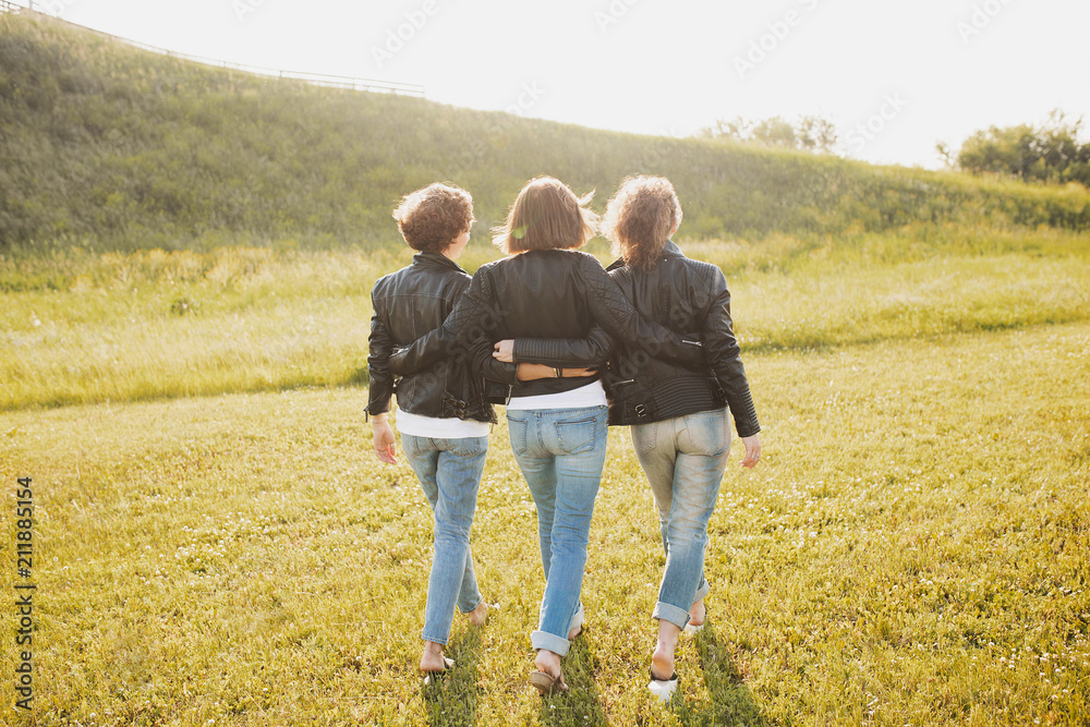 Rear view - three sisters or girlfriends in the same clothes - jeans and leather jackets walking along the lawn of the summer park sunny bright day