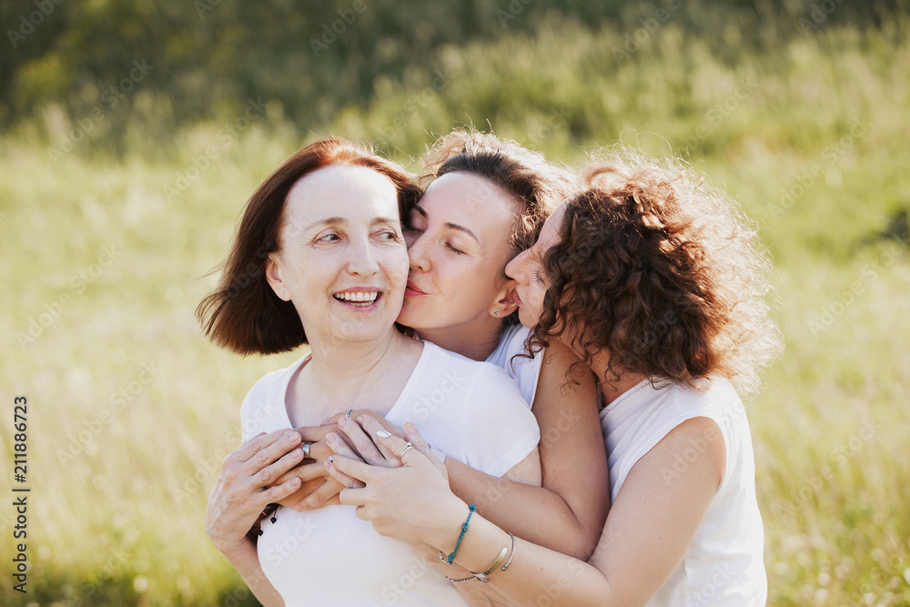 Three pretty women mum and two adult daughters embrace while walking outside the city on a background of a blurred picturesque hill Three pretty women mum and two adult daughters embrace while walking outside the city on a background of a blurred picturesque hill