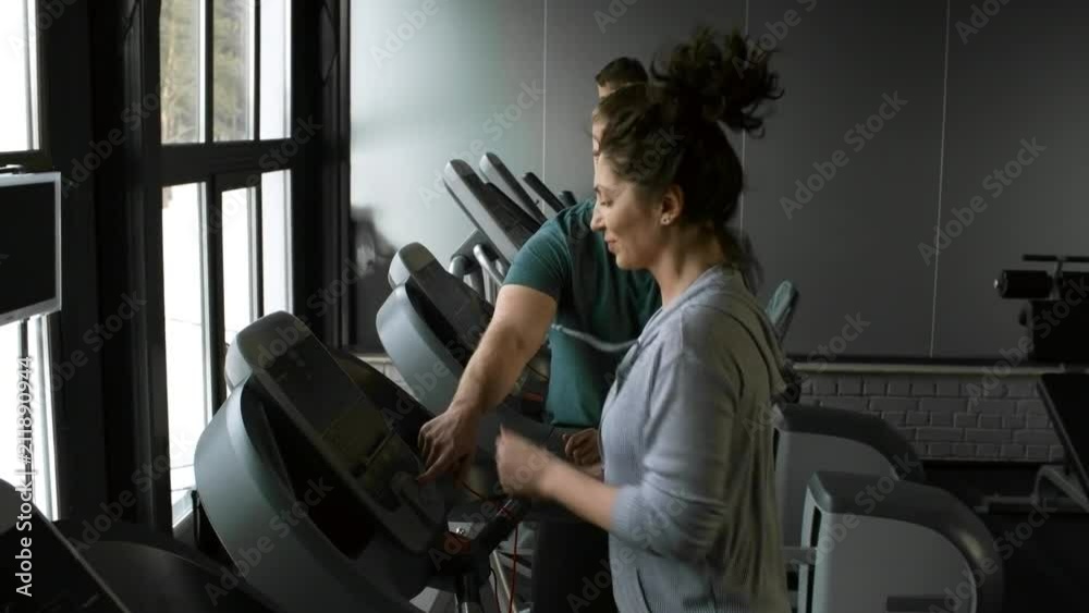 Optimistic woman running on treadmill when working out at the gym, young male coach monitoring her progress and giving thumbs up Optimistic woman running on treadmill when working out at the gym, young male coach monitoring her progress and giving thumbs up