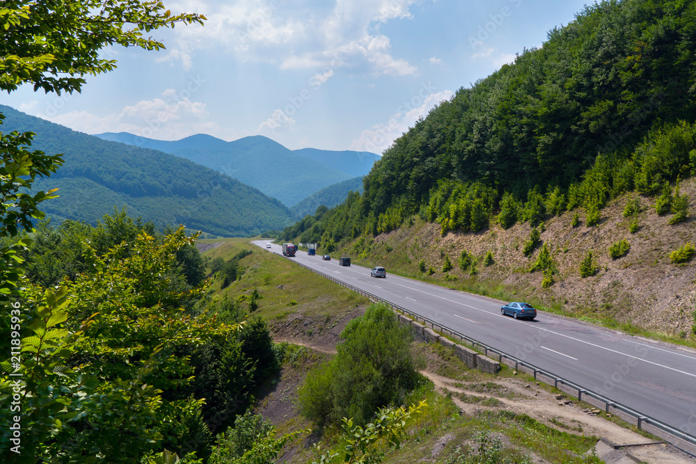 cars driving along a mountain road among forested hills against a blue sky and peaks in the distance cars driving along a mountain road among forested hills against a blue sky and peaks in the distance