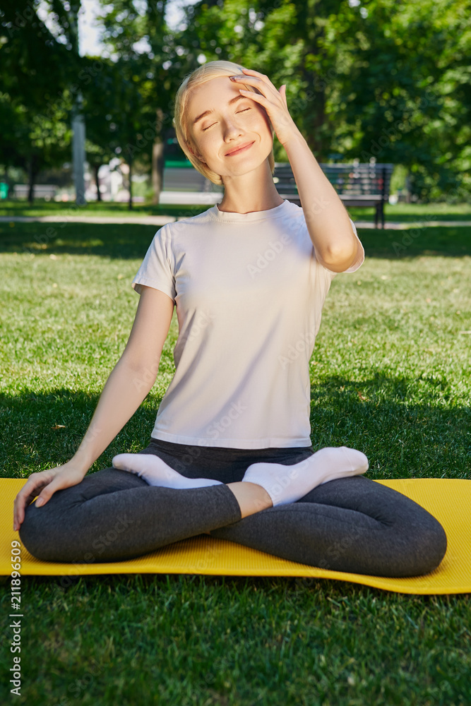 Morning yoga meditation in the summer park. A slim blonde sitting in a  lotus position on an exercise mat, legs crossed. The yogini smiling  happily, touching her forehead, eyes closed. Stock Photo