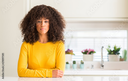 African american woman wearing yellow sweater at kitchen with serious expression on face. Simple and natural looking at the camera.