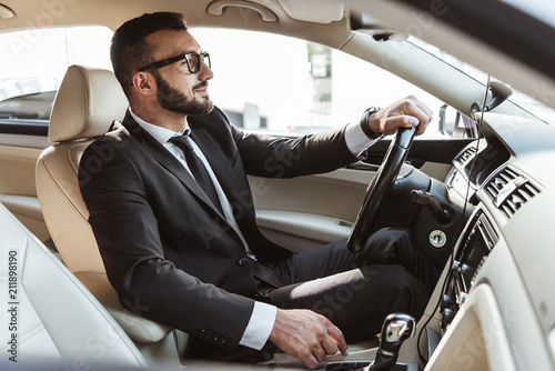 Fotografija side view of handsome driver in suit driving car