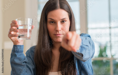 Young woman drinking glass of water at home pointing with finger to the camera and to you, hand sign, positive and confident gesture from the front
