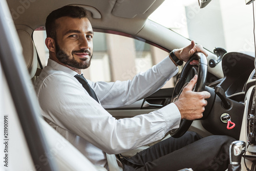 smiling handsome driver holding steering wheel and looking at camera in car