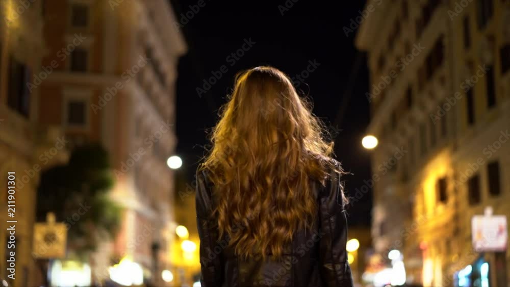Attractive young red hair woman walk away from the camera at night while walking the city streets with lots of lights in the background