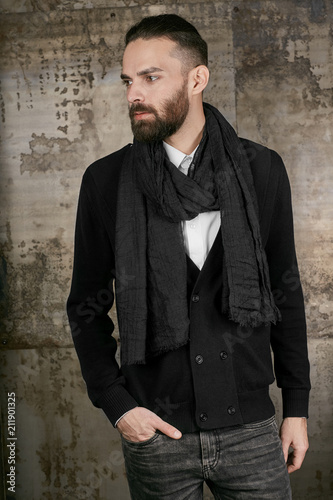 Three quarter isolated studio portrait of a young handsome man in grey jeans, a black cardigan and a scarf. The stylish bearded guy posing over the grunge rusty background, looking to the side.