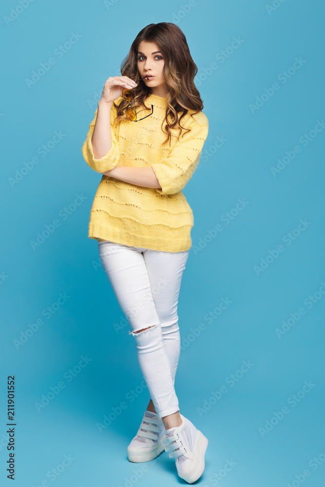 Attractive young woman in yellow sweater posing on blue background. Pretty girl in yellow glasses. Attractive young woman in yellow sweater posing on blue background. Pretty girl in yellow glasses.