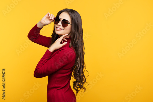 Pretty young lady in red dress and sun glasees is posing with hand on face on orange background.