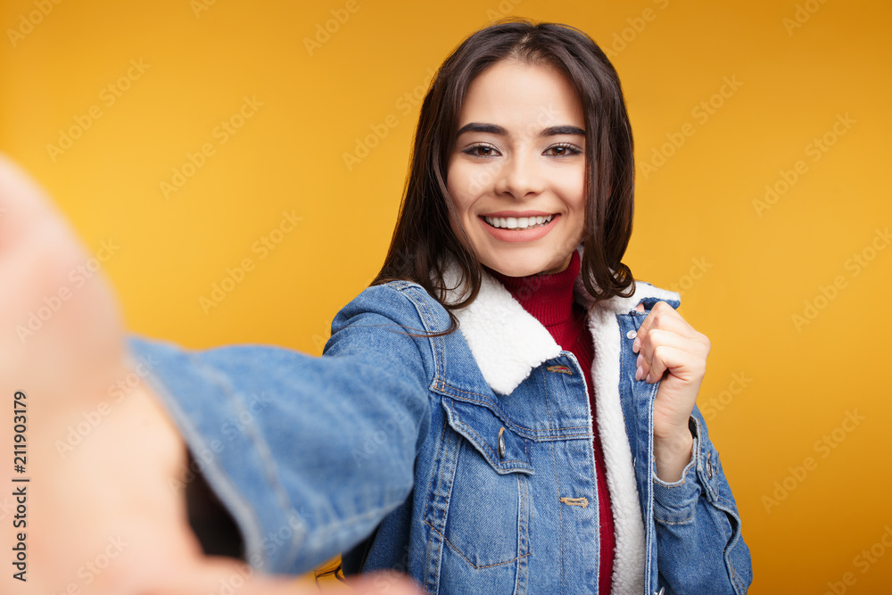 Attractive young lady taking selfie posing in studio in front of orange background. Attractive young lady taking selfie posing in studio in front of orange background.