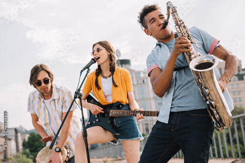 Quadro su tela Multiracial young people with guitar, djembe and saxophone performing on street