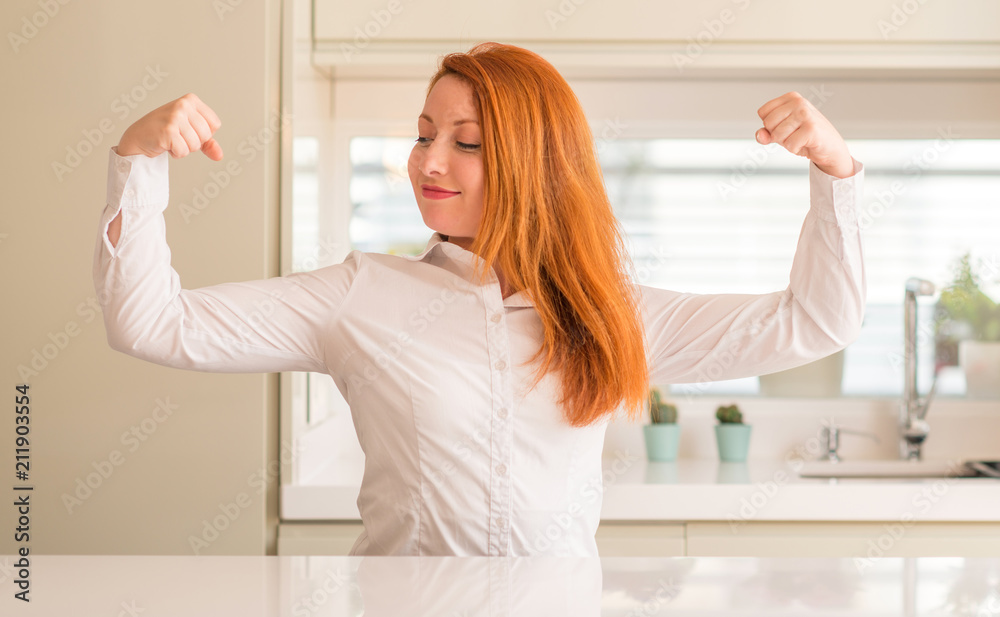 Redhead woman at kitchen showing arms muscles smiling proud. Fitness concept.