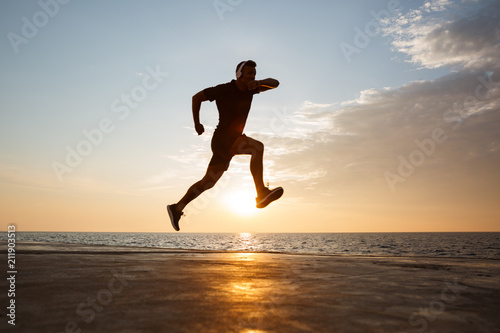 Silhouette of active sporty man 30s jumping and running along pier at seaside  and listening to music via wireless headphones during sunrise
