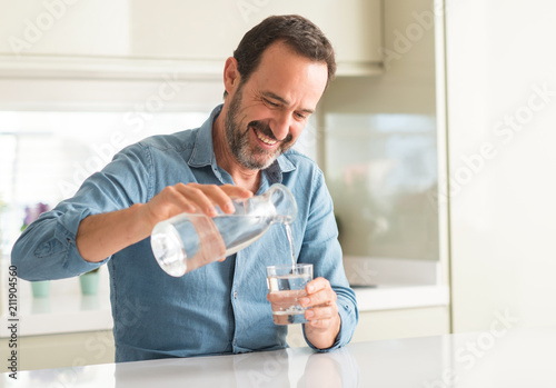 Middle age man drinking a glass of water with a happy face standing and smiling with a confident smile showing teeth